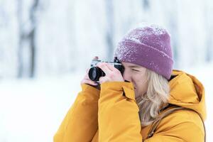 Lycklig flicka fotograf i en gul jacka tar bilder av vinter- i en snöig parkera foto
