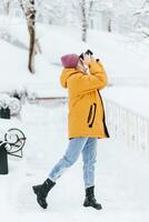 skön flicka i en gul jacka fotograf tar bilder av snö i en vinter- parkera foto
