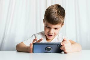 en liten pojke i en vit t-shirt spelar spel på de telefon på Hem. en Lycklig barn utseende på hans smartphone. foto