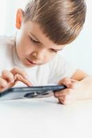 en liten pojke i en vit t-shirt spelar spel på de telefon på Hem. en Lycklig barn utseende på hans smartphone. foto