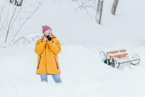 en skön flicka med en kamera i henne händer tar bilder av vinter- i en snöig parkera foto