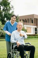 äldre asiatisk senior kvinna på rullstol med asiatisk försiktig vårdgivare och uppmuntra patient, gående i trädgård. med vård från en vårdgivare foto
