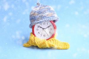 röd rund väckarklocka i stickad ullblå hatt och gul halsduk på blå bakgrund. vintertid koncept. vinter säsong. mysigt och varmt. analog tid 10 10. fallande snö foto