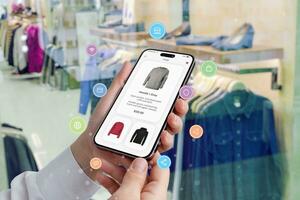 kvinnas händer håll en smartphone med handla app omgiven förbi handla ikoner, medan en boutique ställer ut kläder i de bakgrund foto