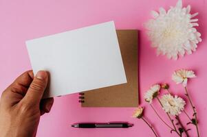hand som håller tomt kort, anteckningsbok, penna och blomma på rosa bakgrund foto