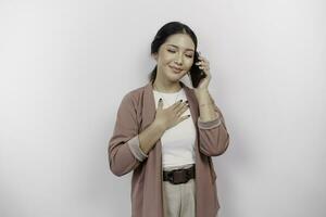 Lycklig uppmärksam tacksam ung asiatisk kvinna anställd bär cardigan och har en telefon ring upp och hand på bröst leende isolerat på vit bakgrund känsla Nej påfrestning, foto