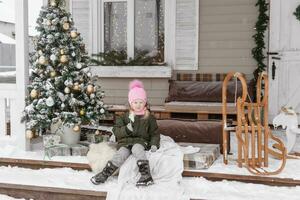 en liten flicka i vinter- kläder är Sammanträde på de veranda av en hus dekorerad för de jul högtider. jul högtider för en barn. foto