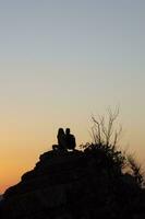 pojke och flicka på en sten tittar på de solnedgång, silhuetter av par tittar på de solnedgång foto