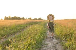 en liten blond flicka i en sugrör hatt promenader i en fält med en bukett av prästkragar. de begrepp av gående i natur, frihet och ett miljövänlig livsstil. foto