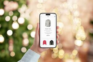 kvinnas hand innehar en mobil telefon med en modern e-handel app, engagerad i jul handla. festlig bokeh av jul träd lampor skapar en Semester handla atmosfär foto