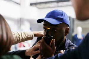 afrikansk amerikan man säkerhet kille använder sig av walkie talkie medan innehav tillbaka folkmassan av shoppare. polis officer hantera stor grupp av människor på Lagra ingång under upptagen Semester handla Seaso foto