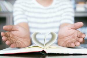 man öppen de hand visa bok med öppen sidor vika ihop en bit av papper hjärta på trä tabell i bibliotek. falsk upp för design aning kärlek till läsa, begrepp utbildning. foto