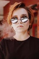 flicka med kort rött hår och spegel solglasögon som röker cigarett