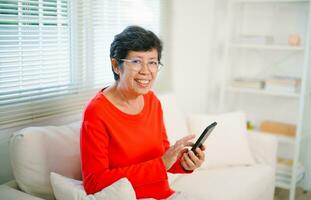Lycklig senior gammal asiatisk kvinna njuter använder sig av mobil appar textning skriver meddelanden sitta på soffa, leende gammal lady innehav smartphone ser smartphone bläddring social media eller inlärning teknologi på Hem foto