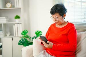 Lycklig senior gammal asiatisk kvinna njuter använder sig av mobil appar textning skriver meddelanden sitta på soffa, leende gammal lady innehav smartphone ser smartphone bläddring social media eller inlärning teknologi på Hem foto