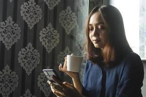 avslappnad asiatisk kvinna som dricker kaffe medan hon använder en mobiltelefon för arbete