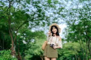 porträtt av asiatisk ung kvinna resande med vävning hatt och korg och en kamera på grön offentlig parkera natur bakgrund. resa resa livsstil, värld resa explorer eller Asien sommar turism begrepp. foto