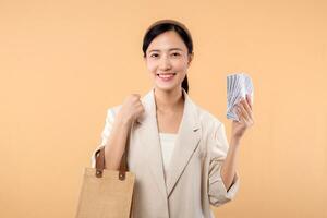 porträtt av Lycklig framgångsrik självsäker ung asiatisk företag kvinna bär vit jacka innehav kontanter pengar dollar och eco väska stående över beige bakgrund. eco vänlig handla begrepp. foto