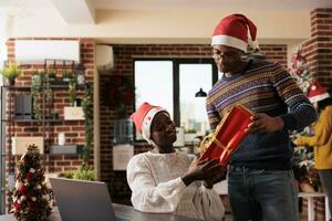 företag anställd ger jul gåva till medarbetare och som visar namn kort bifogad till låda. afrikansk amerikan man och kvinna kollegor utbyte xmas presenterar i festlig dekorerad kontor foto