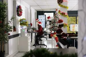 multietniskt företag team bär jul hattar håller på med projekt uppgifter i festlig arbetsplats. mångkulturell anställda kontroll statistisk grafer i dekorerad kontor under xmas säsong foto