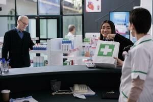 apotek kassör ger klient handla väska med medicin på kolla upp. leende casian kvinna tar inköp paket från farmaceutisk anställd på medicinsk detaljhandeln Lagra disken skrivbord foto