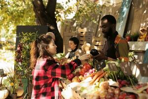 afrikansk amerikan kille Säljare stående Bakom bås försäljning färsk organisk säsong- grönsaker till kvinna kund på lokal- jordbrukare marknadsföra. ung kvinna uppköp naturlig hemodlade producera foto