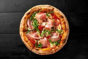 pizza med pelati sås, mozzarella, cabanossi, kyckling filea, bacon, skinka, arugula på svart trä- bakgrund foto