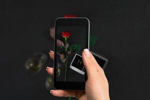 kvinna hand med en smartphone gör en Foto ett mörk röd reste sig och låda med örhängen på svart bakgrund.