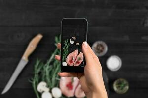 kvinna hand med en smartphone gör en Foto rå kyckling bröst på svart bakgrund, med färsk svamp, vitlök och örter.