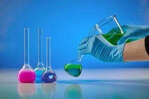 händer i disponibel handskar häller grön flytande från bägare in i medicinsk flaska. lila, rosa kemisk reagenser, blå bakgrund. pandemisk, coronavirus foto