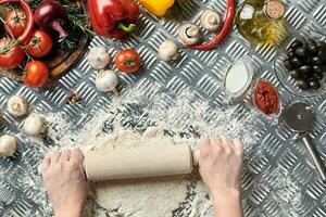 kvinna händer rulla ut deg på metall bakgrund, stänga upp. kock gör deg. tabell med grönsaker och köksutrustning. italiensk kök begrepp. pizza foto