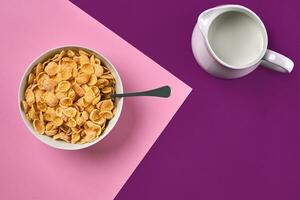 skål med majs flingor, kanna av mjölk och sked på lila och rosa bakgrund, topp se foto