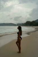 underbar brunett kvinna med perfekt figur Framställ på tropisk strand. bär eleganta svart badkläder. foto