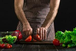 man framställning sallad på en trä- tabell. herr- händer skära de tomat till göra en sallad på svart bakgrund foto