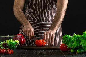 man framställning sallad på en trä- tabell. herr- händer skära de tomat till göra en sallad på svart bakgrund foto