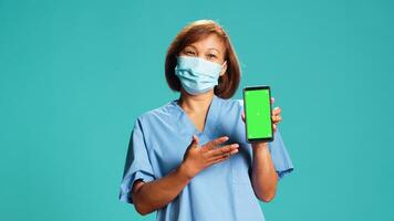 sjuksköterska bär skyddande ansikte mask som visar medicinsk instruktioner video på telefon grön skärm. sjukhus anställd innehav krom nyckel falsk upp smartphone, isolerat över blå studio bakgrund foto