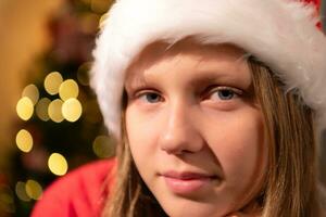 porträtt av Tonårs flicka i santa hatt Sammanträde på stol i främre av jul träd, glad jul och Lycklig högtider foto