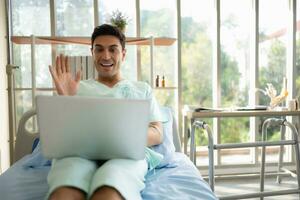 Lycklig ung man använder sig av bärbar dator dator medan Sammanträde på säng i sjukhus avdelning foto