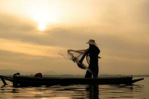 silhuett av fiskare på soluppgång, stående ombord en rodd båt och gjutning en netto till fånga fisk för mat foto