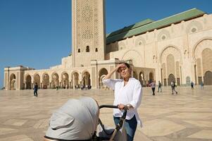 charmig kvinna turist, ung mor tryckande bebis pråm, besöker de hassan ii moské i casablanca. foto