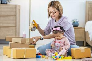 ung asiatisk mor är förpackning de handelsvaror produkt paket medan arbetssätt på Hem redo till leverera medan henne litet barn unge är spelar runt om med henne leksak foto