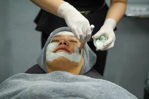 närbild kosmetolog är applicering ansiktsbehandling vit mask grädde på kvinna klient ansikte i skönhet klinik. foto