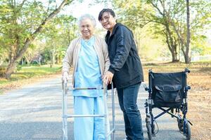 vårdgivare hjälp asiatisk senior kvinna promenad med rollator med rullstol på parkera. foto