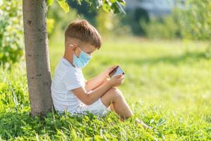 en unge i en medicinsk mask sitter på de gräs och utseende i de telefon tecknade serier i de sommar på solnedgång. barn med en mobil telefon i hans händer. förebyggande mot coronavirus covid-19 under en pandemi foto