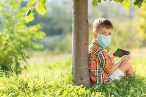 en unge i en medicinsk mask sitter på de gräs och utseende i de telefon tecknade serier i de sommar på solnedgång. barn med en mobil telefon i hans händer. förebyggande mot coronavirus covid-19 under en pandemi foto