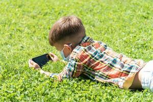 en barn i en medicinsk mask lögner på de gräs och utseende i de telefon tecknade serier i de sommar på solnedgång. unge med en mobil telefon i hans händer. förebyggande mot coronavirus covid-19 under en pandemi foto