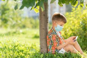 en unge i en medicinsk mask sitter på de gräs och utseende i de telefon tecknade serier i de sommar på solnedgång. barn med en mobil telefon i hans händer. förebyggande mot coronavirus covid-19 under en pandemi foto