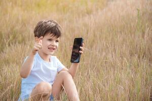 en skön barn Sammanträde på de gräs talar förbi telefon i de sommar på solnedgång. pojke kommunicerar på mobil foto