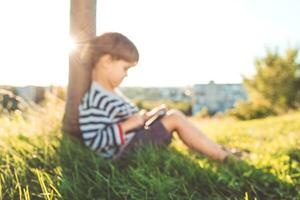 en skön barn Sammanträde på de gräs spelar på de telefon i en spel i de sommar på solnedgång. pojke har roligt i natur foto