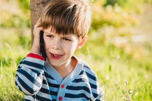en skön barn Sammanträde på de gräs talar förbi telefon i de sommar på solnedgång. pojke kommunicerar på mobil foto